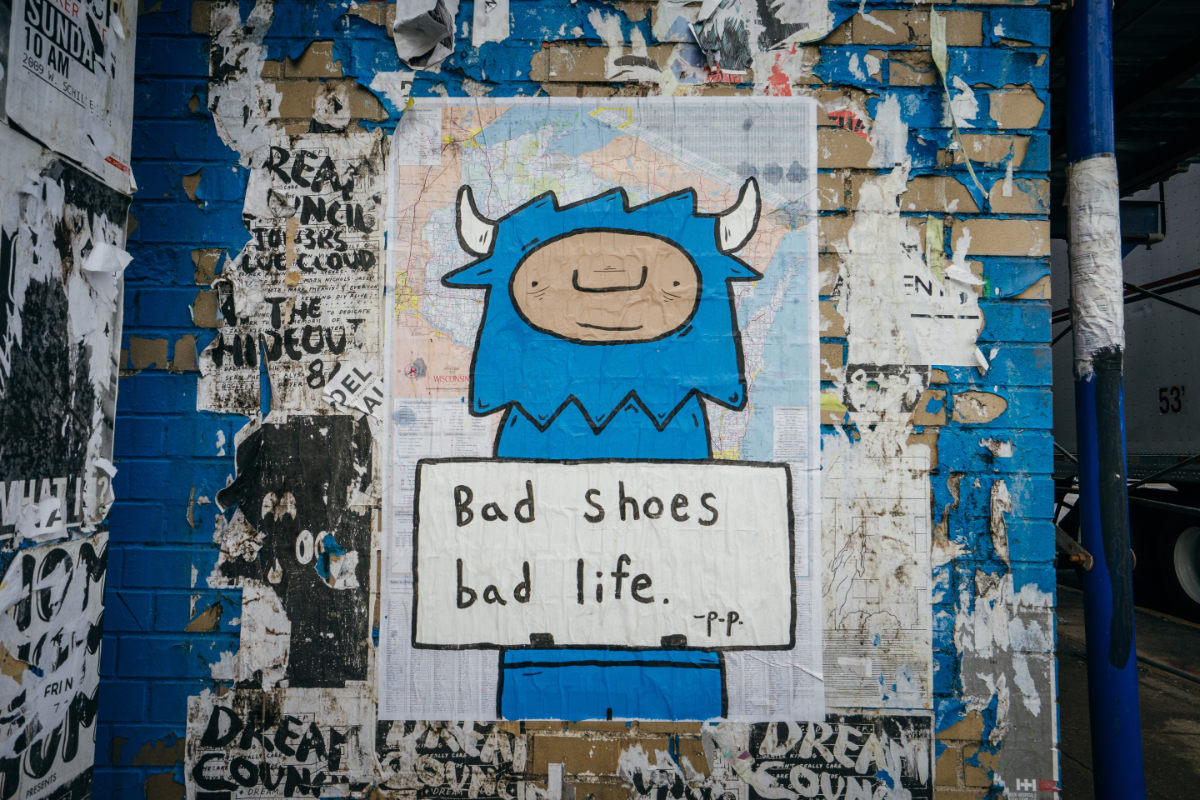 Photo of a graffitied charatcter on a messy brick wall. The character is holding a sign that says, "Bad Shoes, bad life."